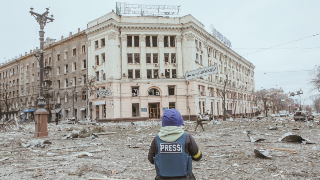 Journalist in front of bombed building in Ukraine.
